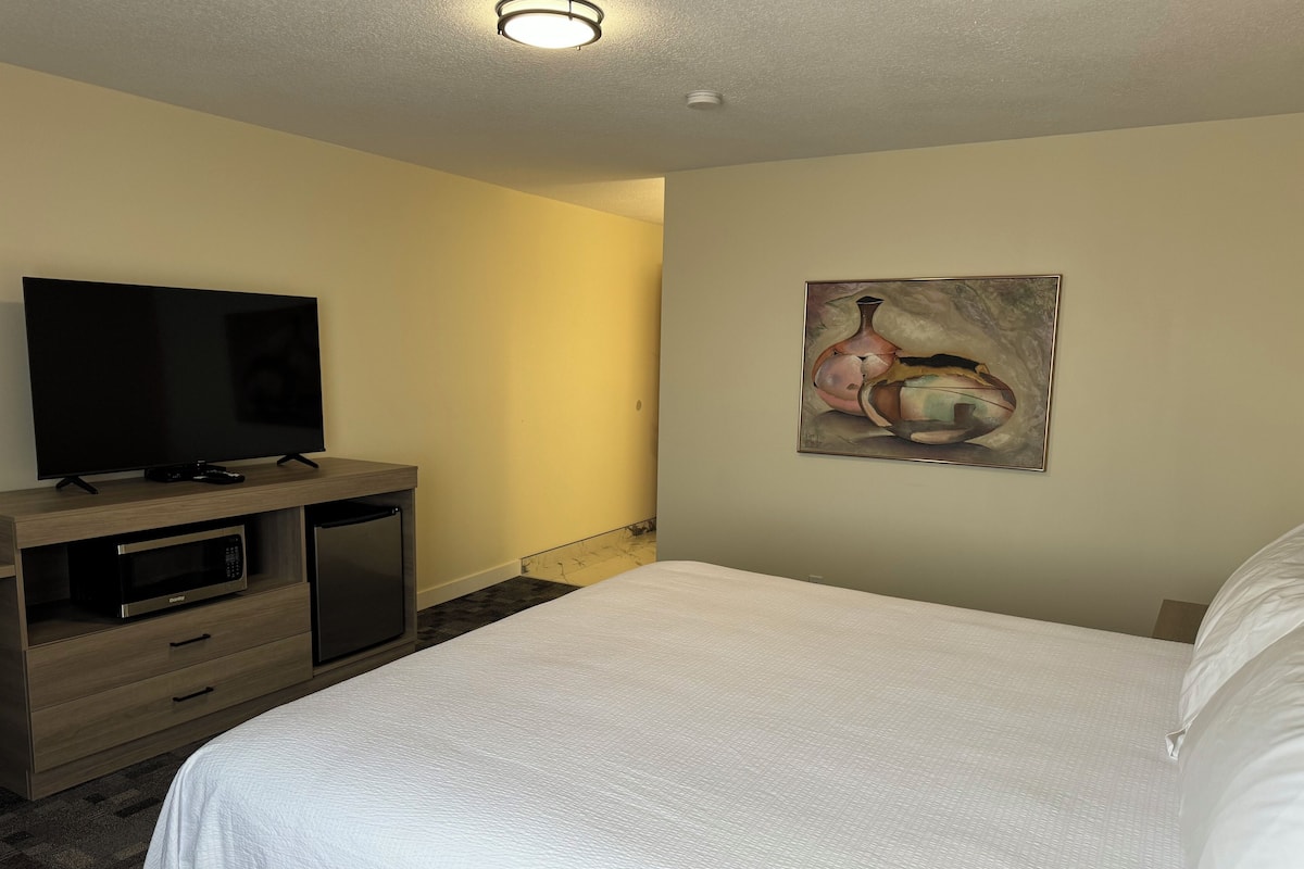 A hotel room with a bed, a flat-screen TV on a wooden console, a microwave, and a painting on the wall.