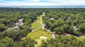 Vista aérea de um campo de golfe com um obstáculo aquático, rodeado por árvores densas e propriedades residenciais.