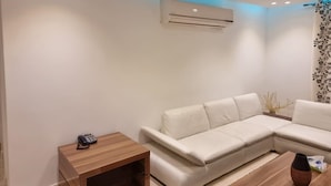 A modern living room with a white sectional sofa, a wooden side table with a phone, and a small vase on a wooden coffee table. - Rehab City- Appartement Cosy-chic Près de Toutes Commodités (Cairo Governorate)