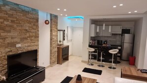 A modern living room with a stone wall, a flat-screen TV, a wooden coffee table, and a kitchen area with bar stools and a refrigerator. - Rehab City- Appartement Cosy-chic Près de Toutes Commodités (Cairo Governorate)