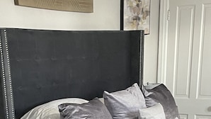 A bedroom with a bed, grey and white bedding, a dark headboard, and two white planters with green plants on the floor. - Harrisburg apt Great River View (Harrisburg)