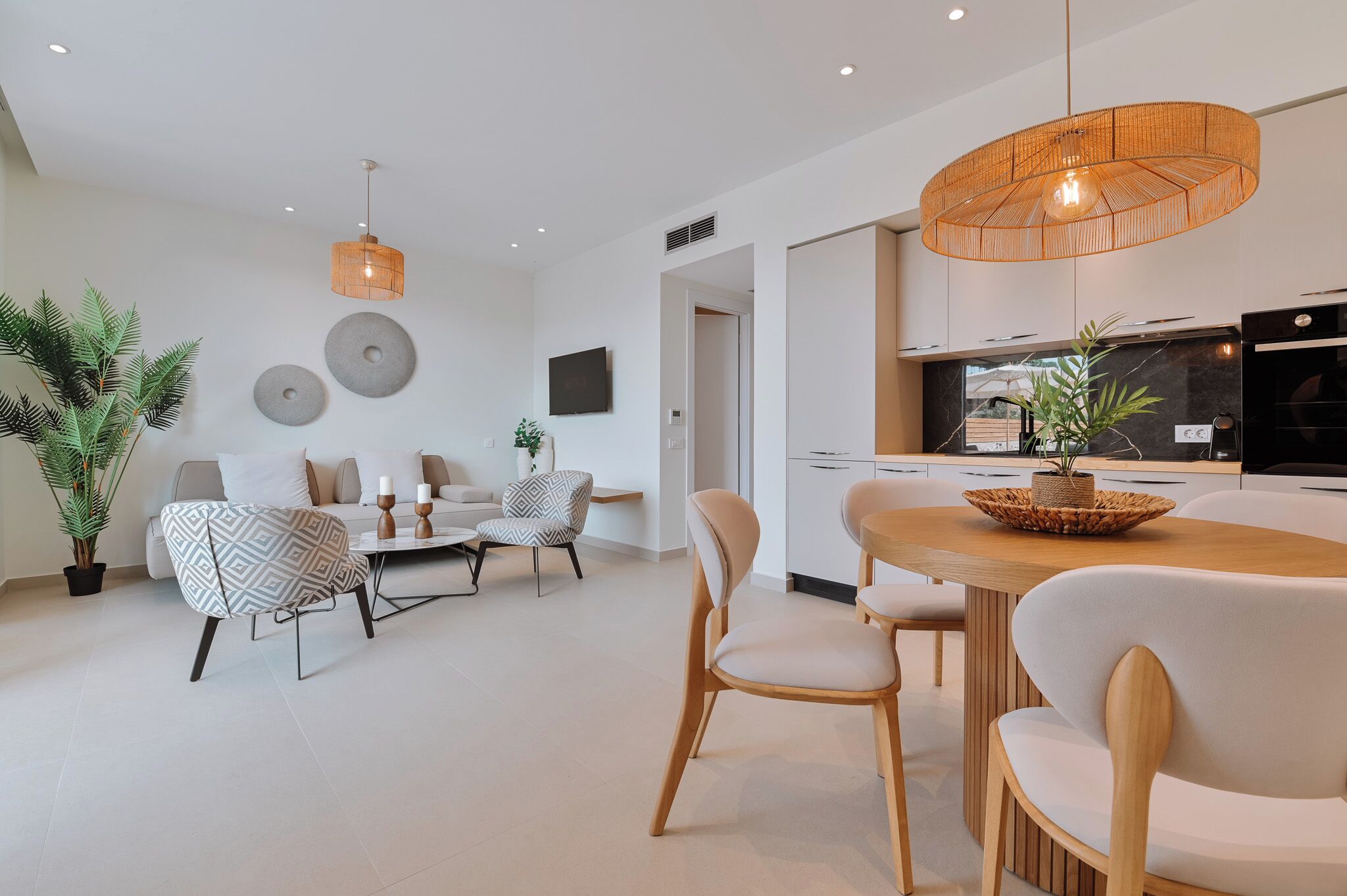 A modern living room with a dining area, featuring a round wooden table, beige chairs, and a hanging light fixture.