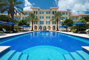 Una amplia piscina con azulejos azules, rodeada de sillones de descanso y sombrillas, con un edificio grande con múltiples ventanas y balcones al fondo.