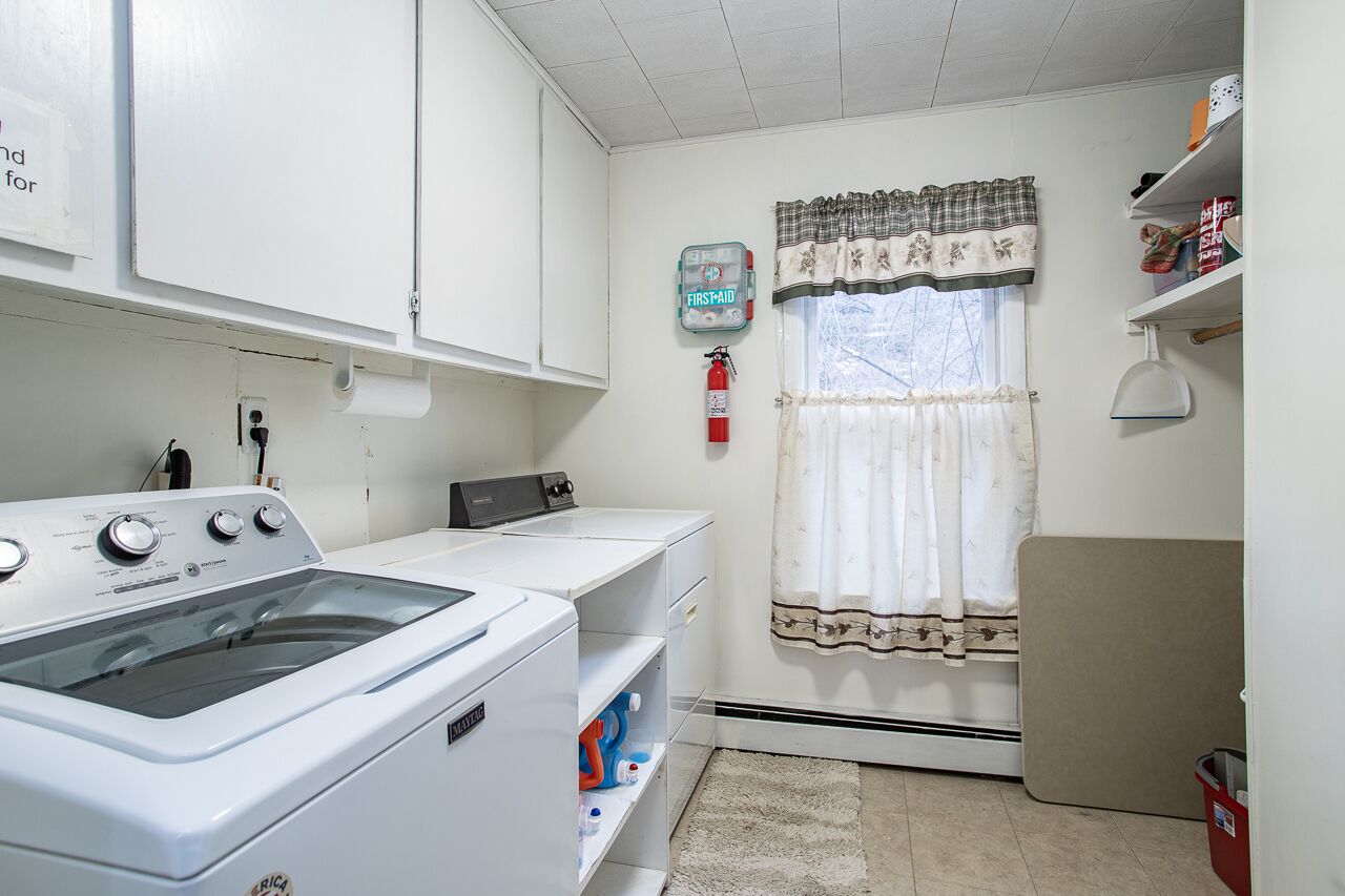 A laundry room with a front-loading washing machine, a dryer, a shelf with cleaning supplies, a fire extinguisher, and a window with curtains.