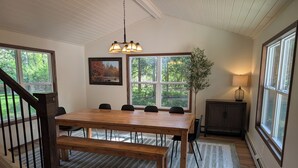Un comedor con mesa de madera, sillas, un banco, una lámpara de araña, un cuadro, una planta en maceta y un aparador.