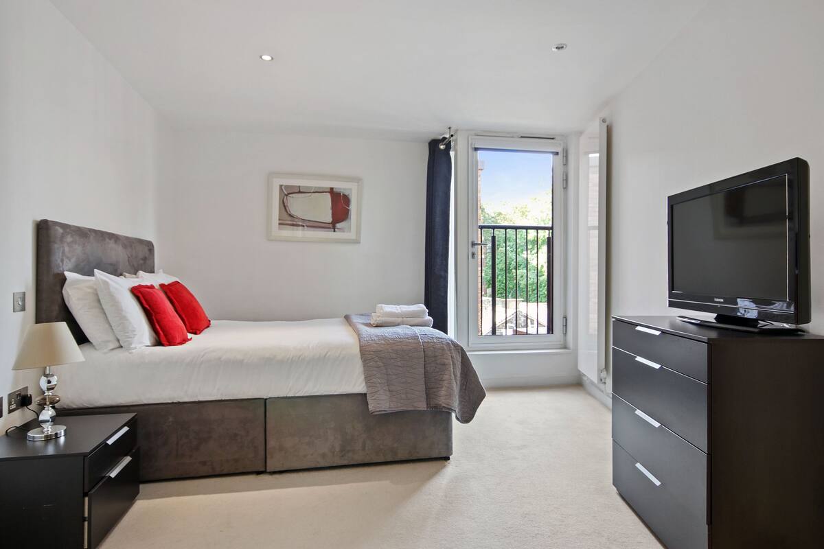 A modern bedroom with a bed, a dresser, a television, and a window with a view of greenery.