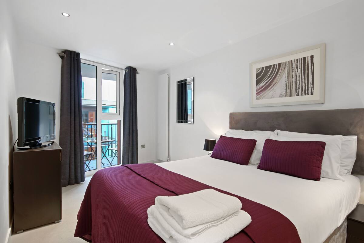 A hotel room with a large bed, a television on a cabinet, a balcony with a view, and a painting on the wall.