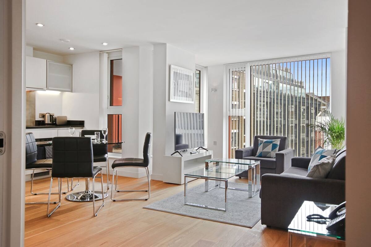 A modern living room with a glass coffee table, a sofa, and a dining area with a kitchen in the background.