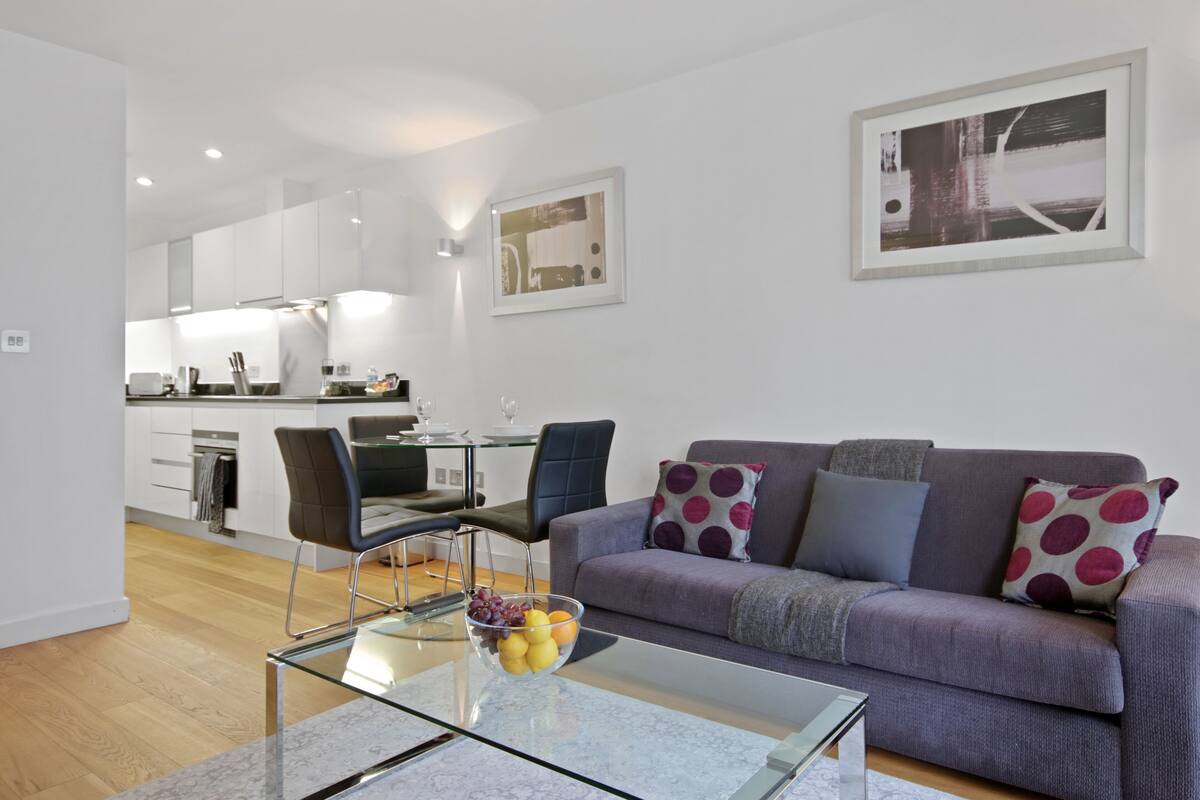 A modern living room with a glass coffee table, a purple sofa with patterned cushions, and a dining area with a glass table and chairs.