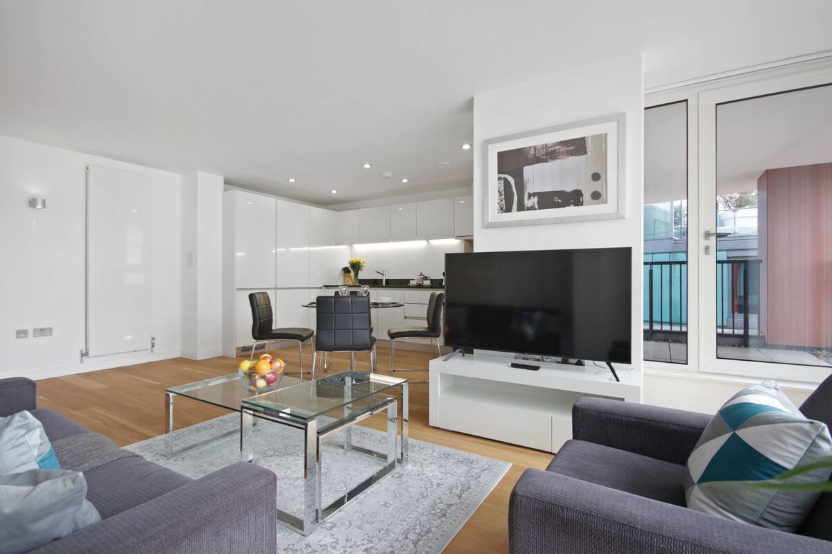A modern living room with a glass coffee table, grey sofas, a flat-screen TV, and a kitchen in the background.