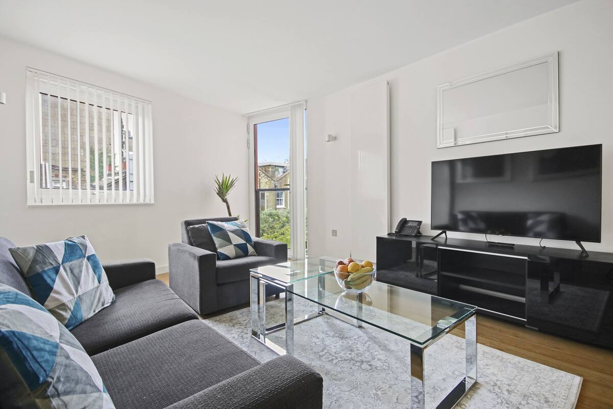 A modern living room with a sofa, a glass coffee table, a flat-screen TV, and a balcony with a view.