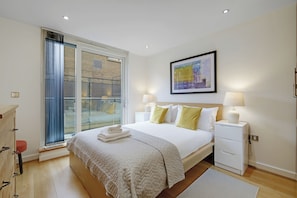 Um quarto com cama, mesas de cabeceira, uma janela com persianas, um quadro na parede e um banquinho vermelho.