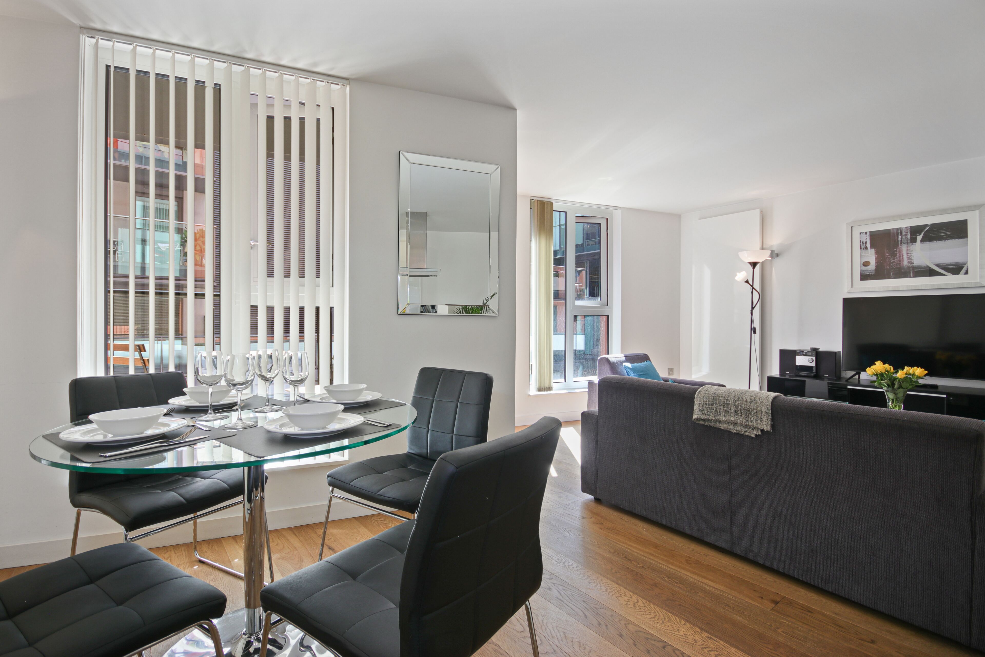 A modern living room with a glass dining table, black chairs, a grey sofa, and a flat-screen TV.