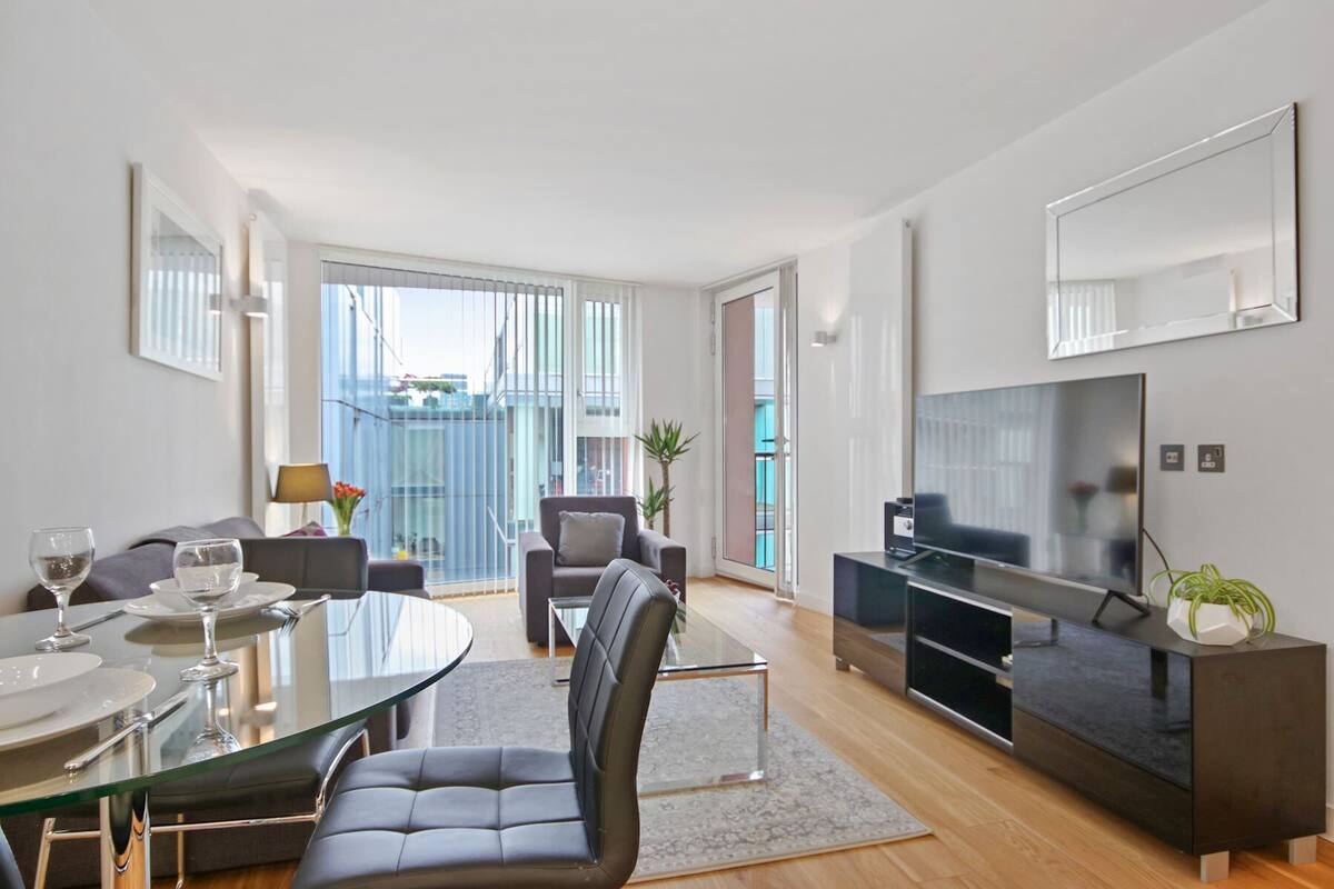 A modern living room with a glass dining table, comfortable seating, a flat-screen TV, and a balcony with a view.