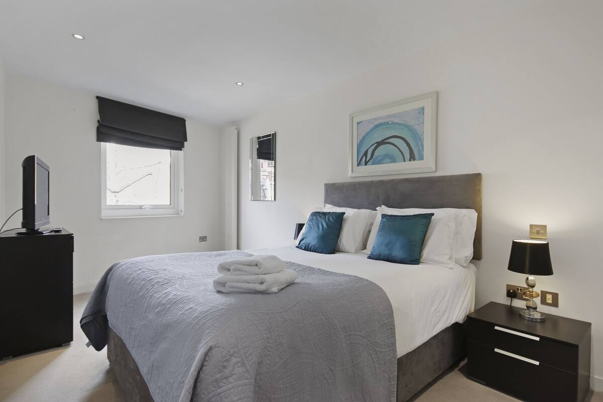A modern bedroom with a large bed, a nightstand with a lamp, a TV on a stand, and a window with a view.
