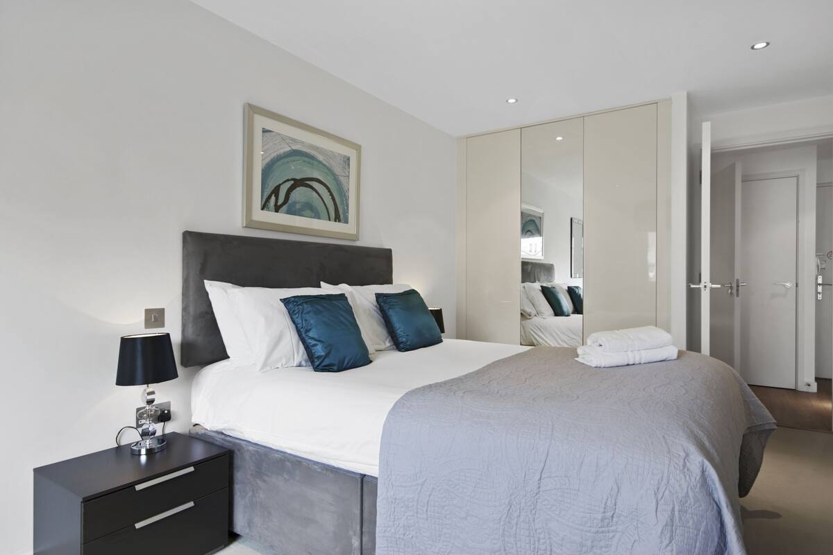 A modern bedroom with a large bed, a bedside table with a lamp, a framed artwork on the wall, and a wardrobe with a mirror.