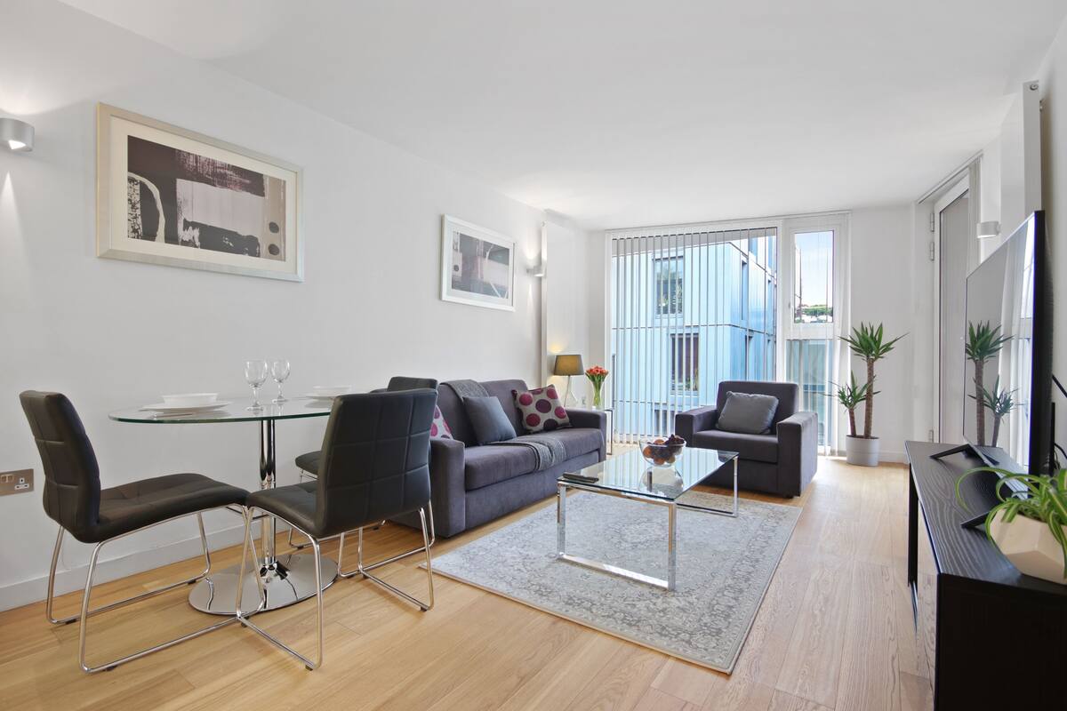 A modern living room with a glass dining table, black chairs, a grey sofa, and a black TV stand.