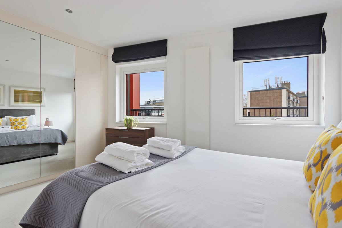 A modern hotel room with a large bed, a wardrobe, a window with a view of buildings, and a small table with a vase.