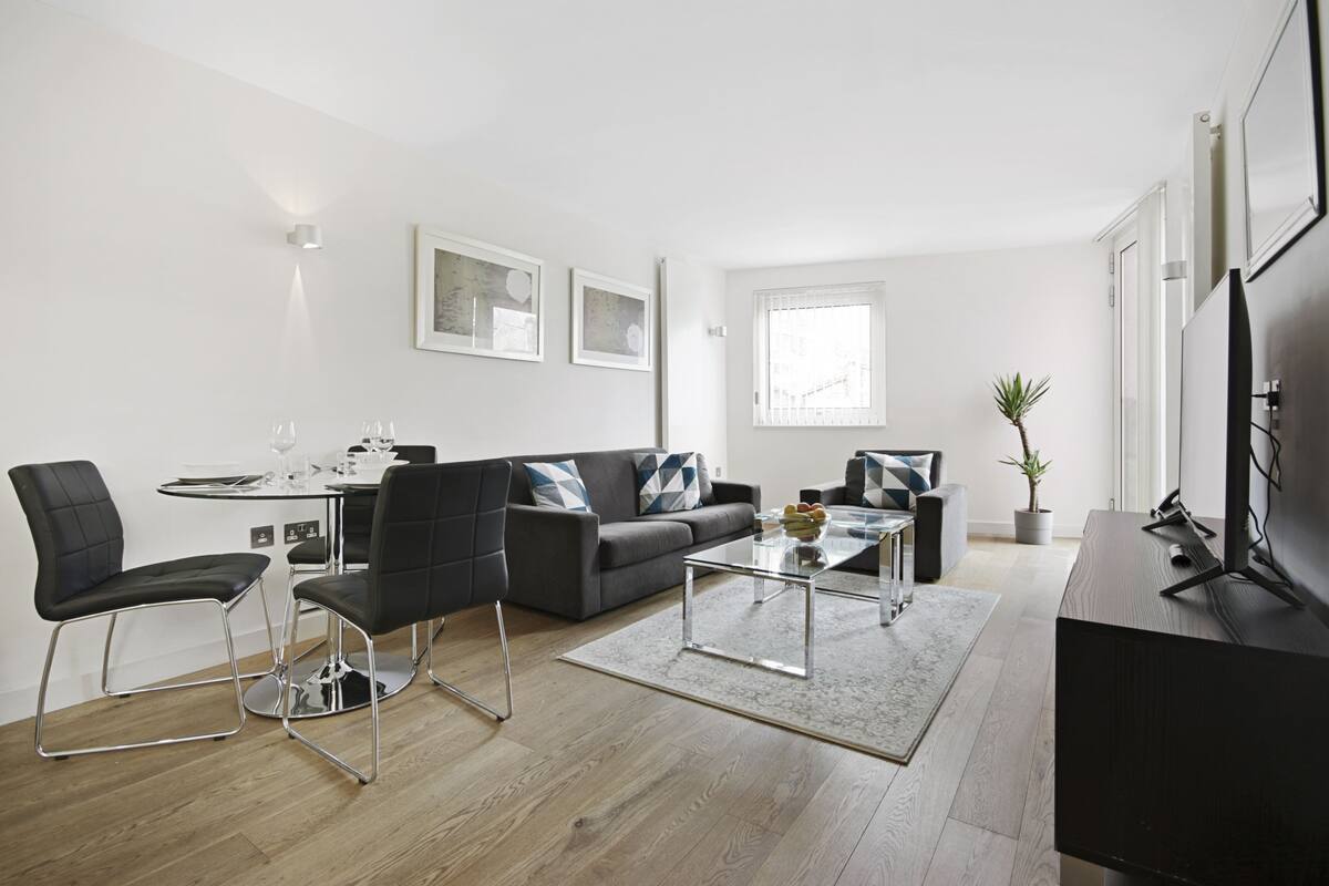 A modern living room with a sofa, coffee table, and chairs. There is a dining table with chairs, a TV, and a window with blinds.