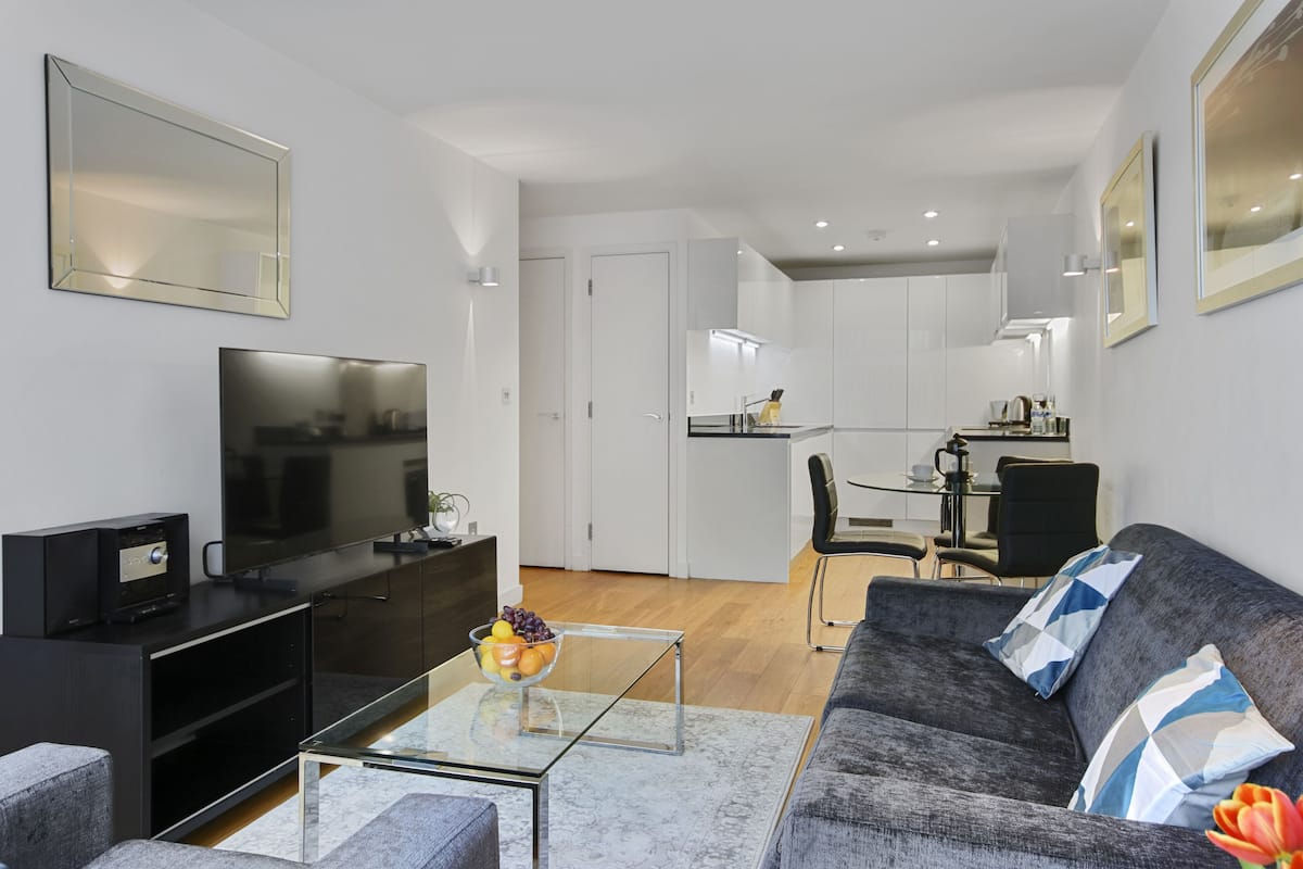 A modern living room with a flat-screen TV, a glass coffee table, and a grey sofa with patterned cushions.
