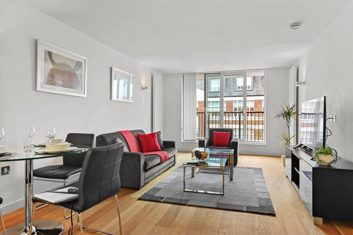A modern living room with a glass dining table, black leather sofa, and a large window with a view of buildings.