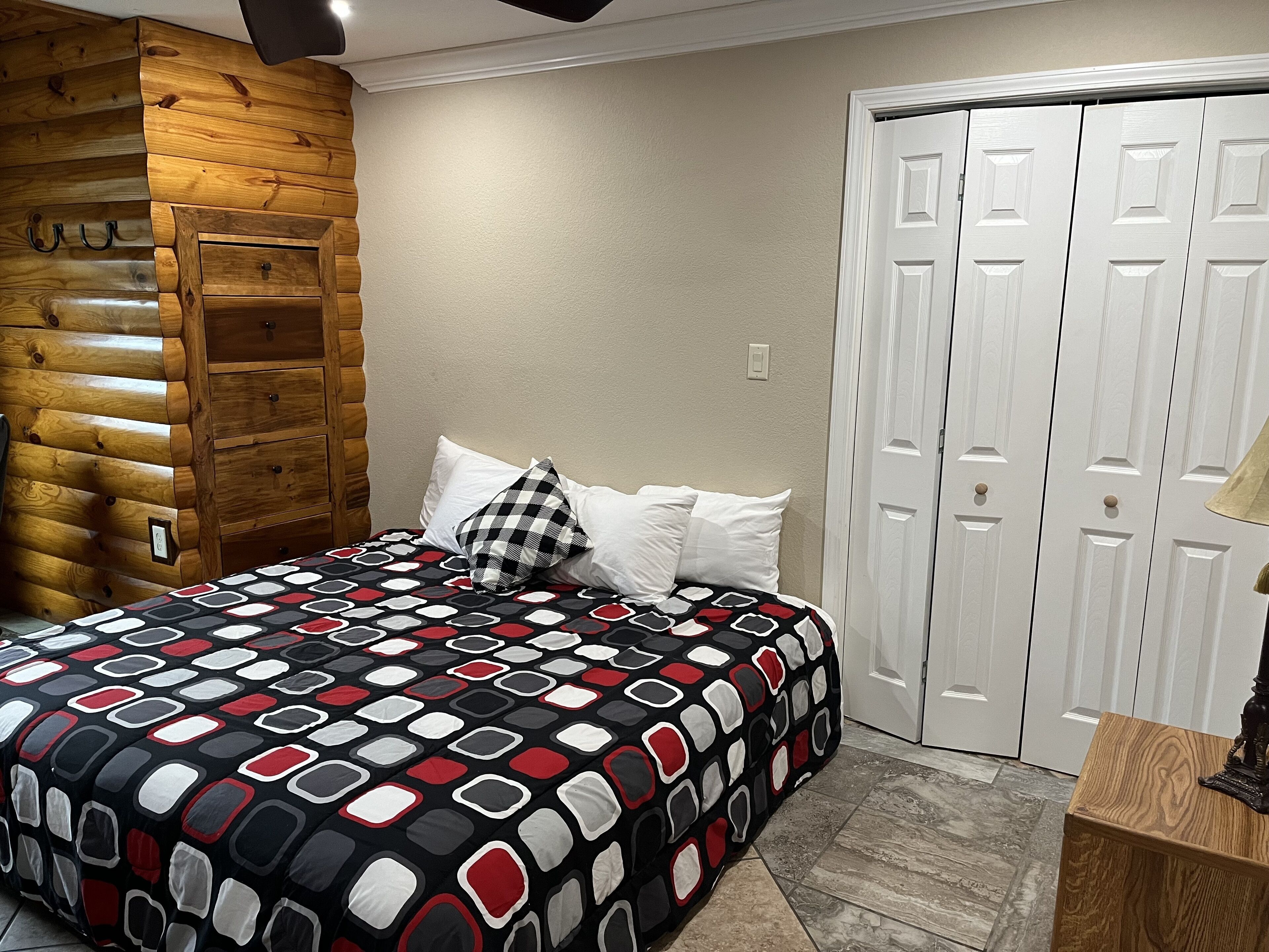 Een slaapkamer met een houten wand van boomstammen, een bed met een beddengoed met een patroon, witte kussens en een witte deur.