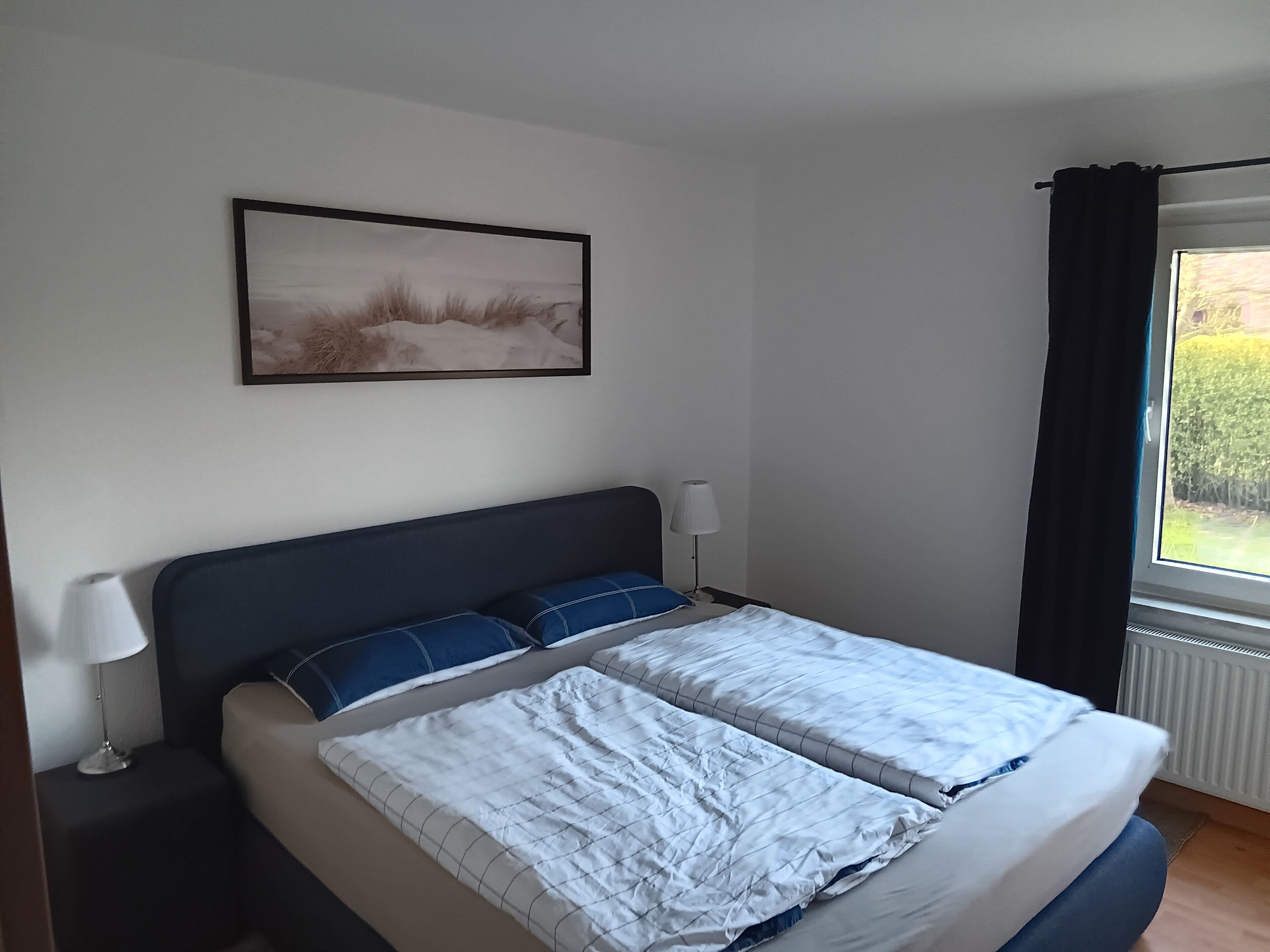 A bedroom with a bed, a bedside table with a lamp, a window with curtains, and a painting on the wall.