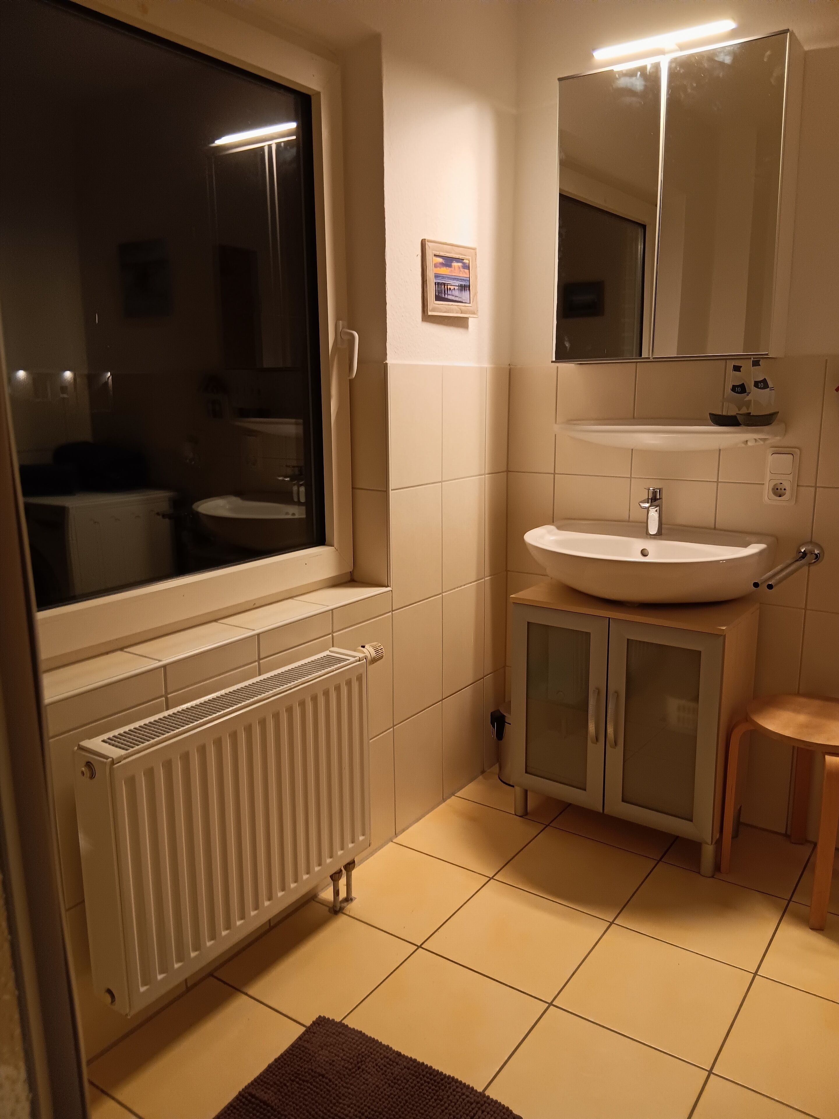 A bathroom with a window, a radiator, a sink, a mirror, and a cabinet.