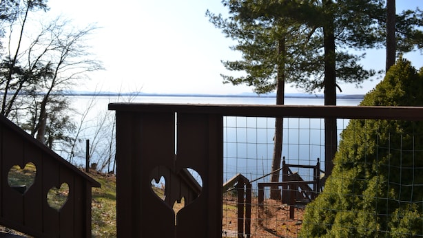 A wooden fence with heart-shaped cutouts, a wire mesh gate, and a view of a body of water and trees in the background. - Lake Champlain house: sunsets, mountains, fishing (Georgia)