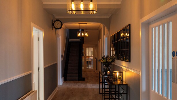 A hallway with a staircase, a clock, and a bar area with a vase of flowers. - The Crossing (Kingussie)