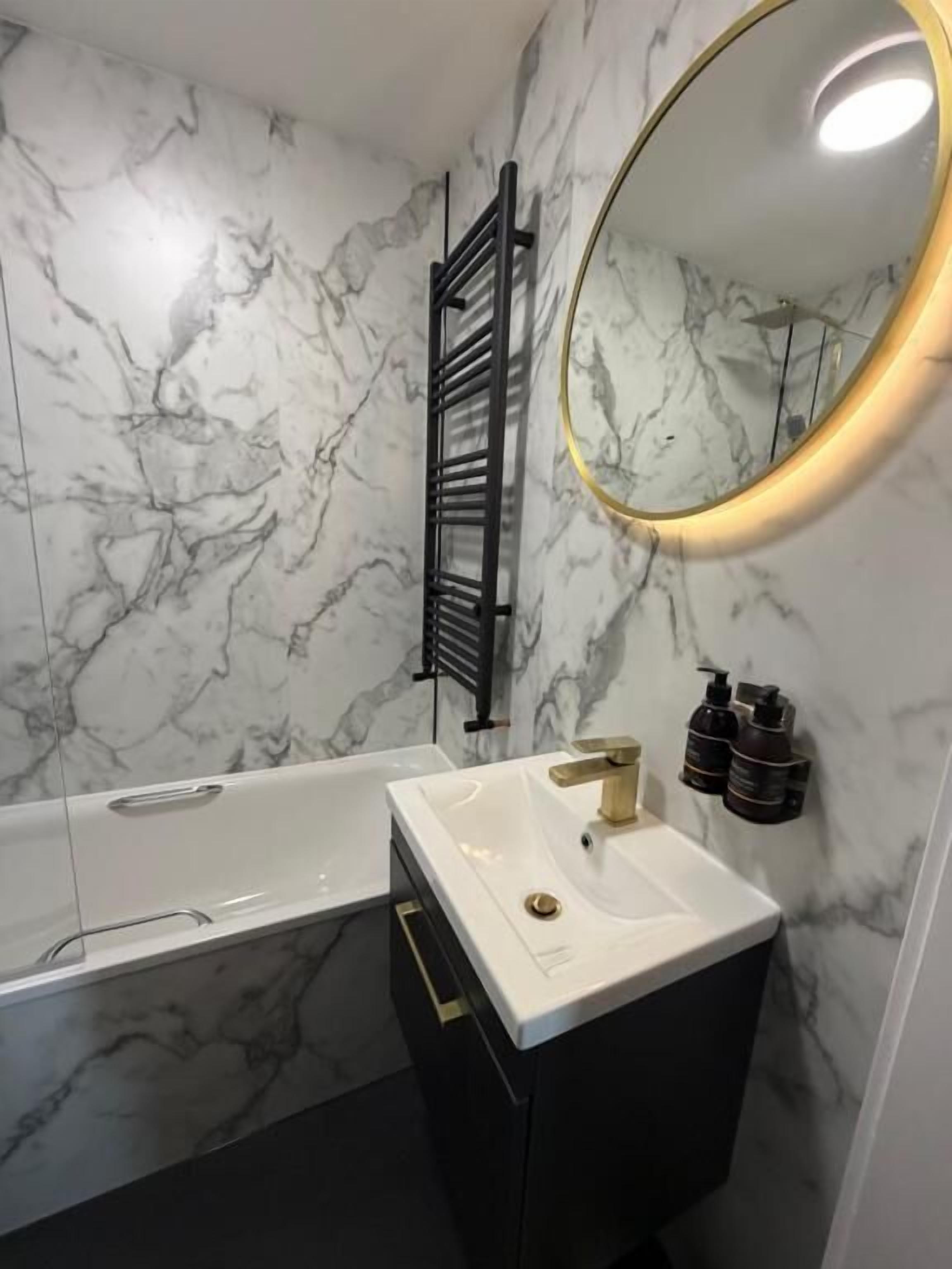 A bathroom with a white sink, black vanity, marble walls, a black towel rack, and a round mirror with a gold frame.