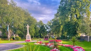 A landscaped garden with a central monument, surrounded by vibrant flower beds and a palm tree. - The Crossing (Kingussie)