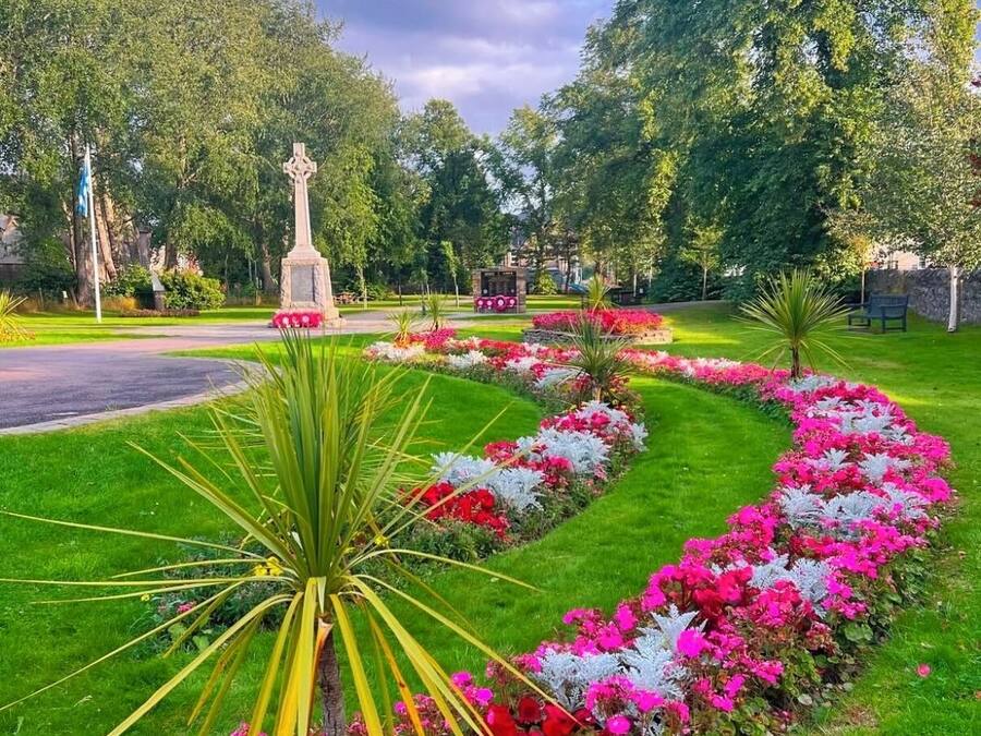 Jardín ajardinado con un monumento central, rodeado de coloridos macizos de flores y una palmera.