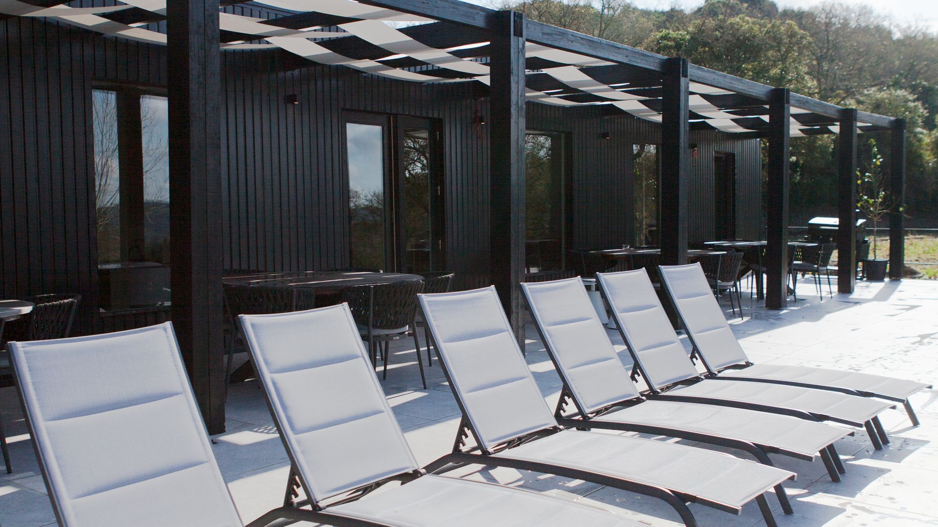 A covered outdoor seating area with white cushioned chairs and tables, a wooden pergola structure, and a building with large windows in the background.