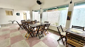 Un'ampia sala da pranzo interna con tavoli e sedie in legno, ampie finestre con vista sul verde e un ventilatore a soffitto.