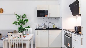 Cocina compacta con armarios blancos, un microondas negro, un lavamanos y una estufa. Hay una mesa de comedor con sillas y una pequeña planta en maceta.