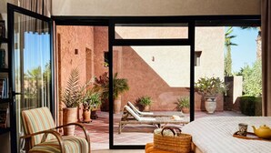 A spacious room with a large bed, a striped armchair, a wicker basket, and a view of an outdoor patio with lounge chairs and potted plants. - Villa Baidane Avec sa Somptueuse vue sur L'atlas (sidi abd alla ghiat)