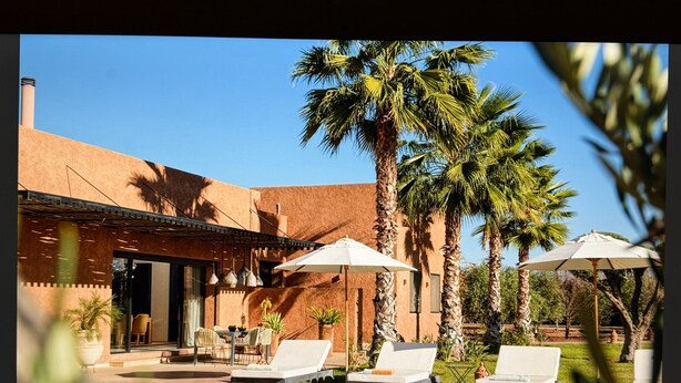 A poolside area with lounge chairs and umbrellas, surrounded by palm trees and a building with a brick facade. - Villa Baidane Avec sa Somptueuse vue sur L'atlas (sidi abd alla ghiat)