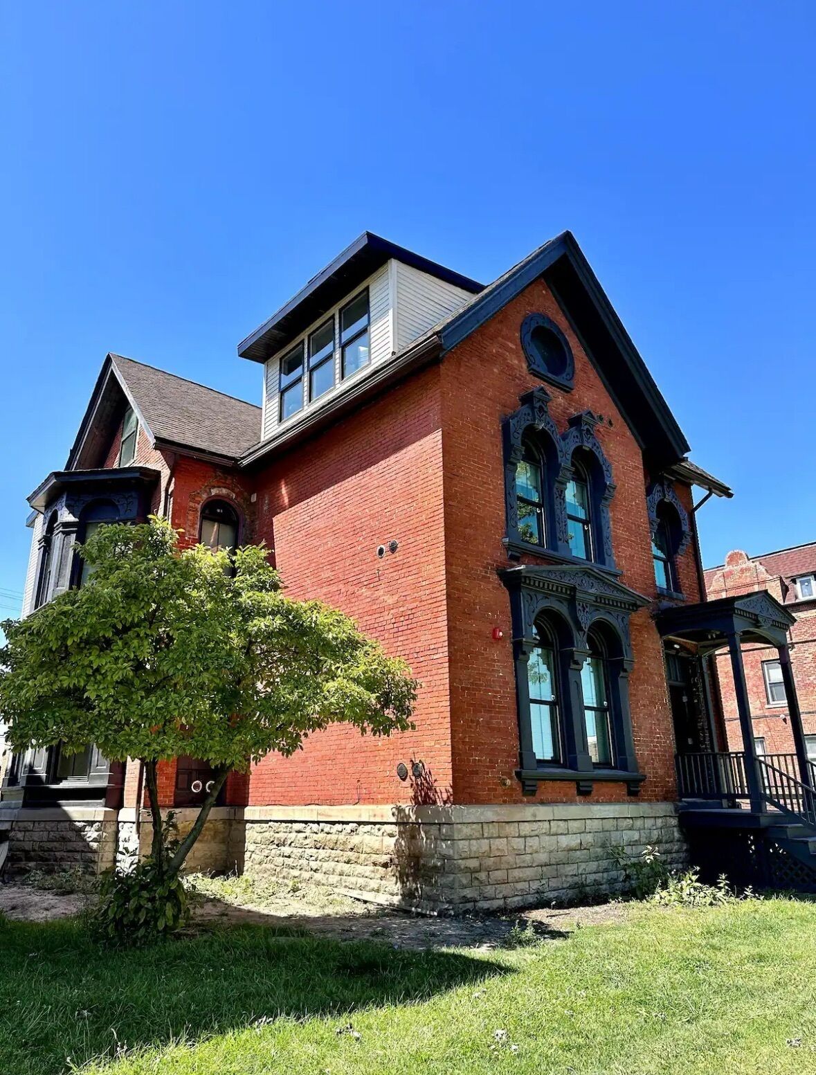 A red brick building with a gabled roof and a large tree in the front yard.