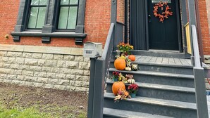 Un bâtiment en briques, agrémenté de décorations noires, avec une porte d’entrée noire ornée d’une guirlande, et un petit porche décoré de citrouilles et de fleurs.