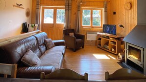 L’intérieur chaleureux d’un chalet avec un plafond en bois, un canapé en cuir, une chaise en bois, un téléviseur et un poêle à bois.