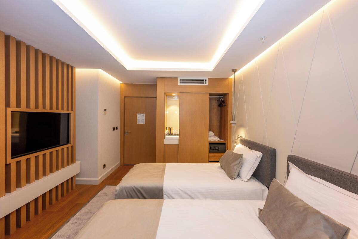 Habitación de hotel moderna con dos camas, un televisor de pantalla plana montado en un panel de madera, vestidor y techo con iluminación empotrada.