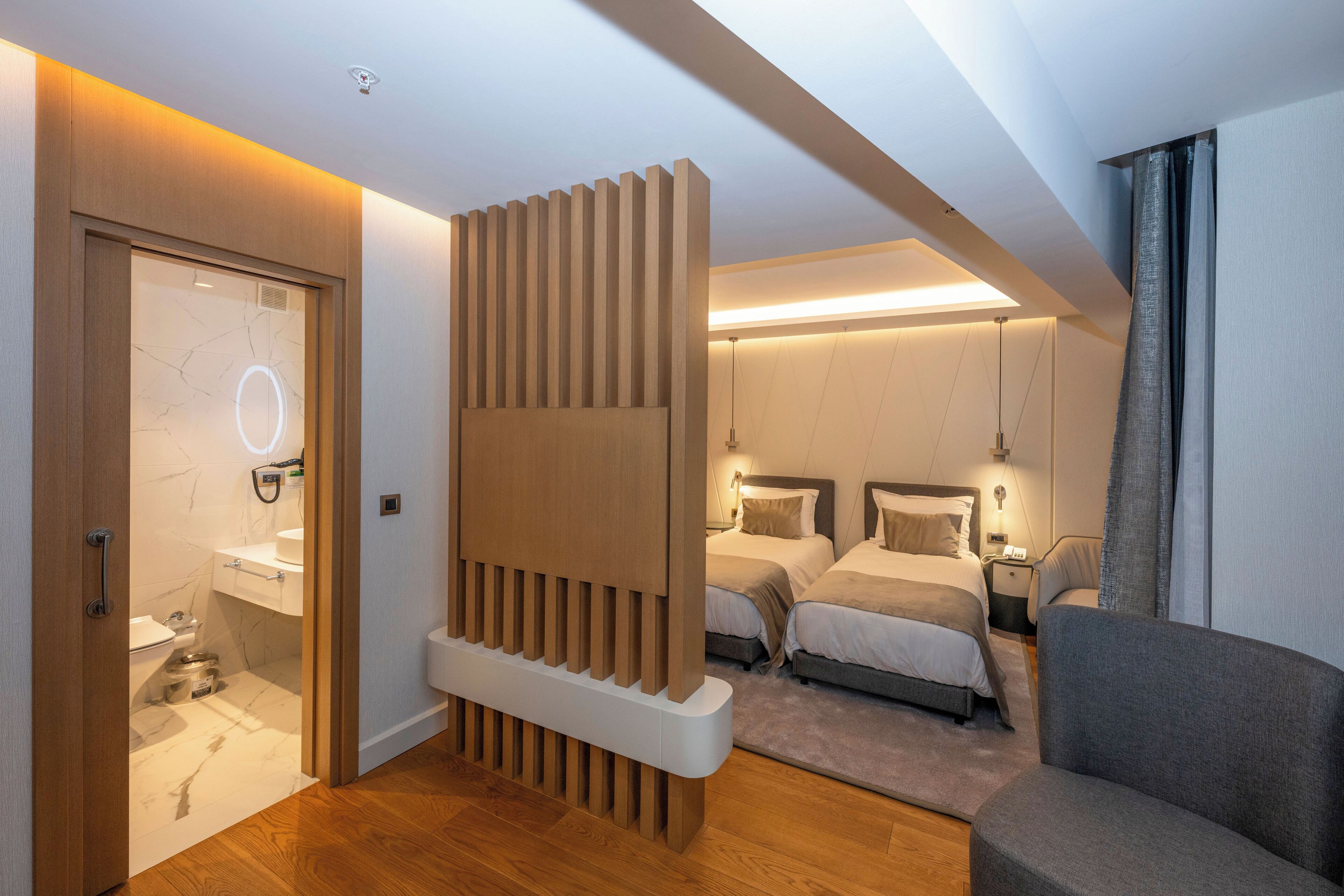 Una habitación de hotel moderna con un separador de madera, dos camas, un sofá, un baño con lavamanos de mármol y una lámpara en el techo.