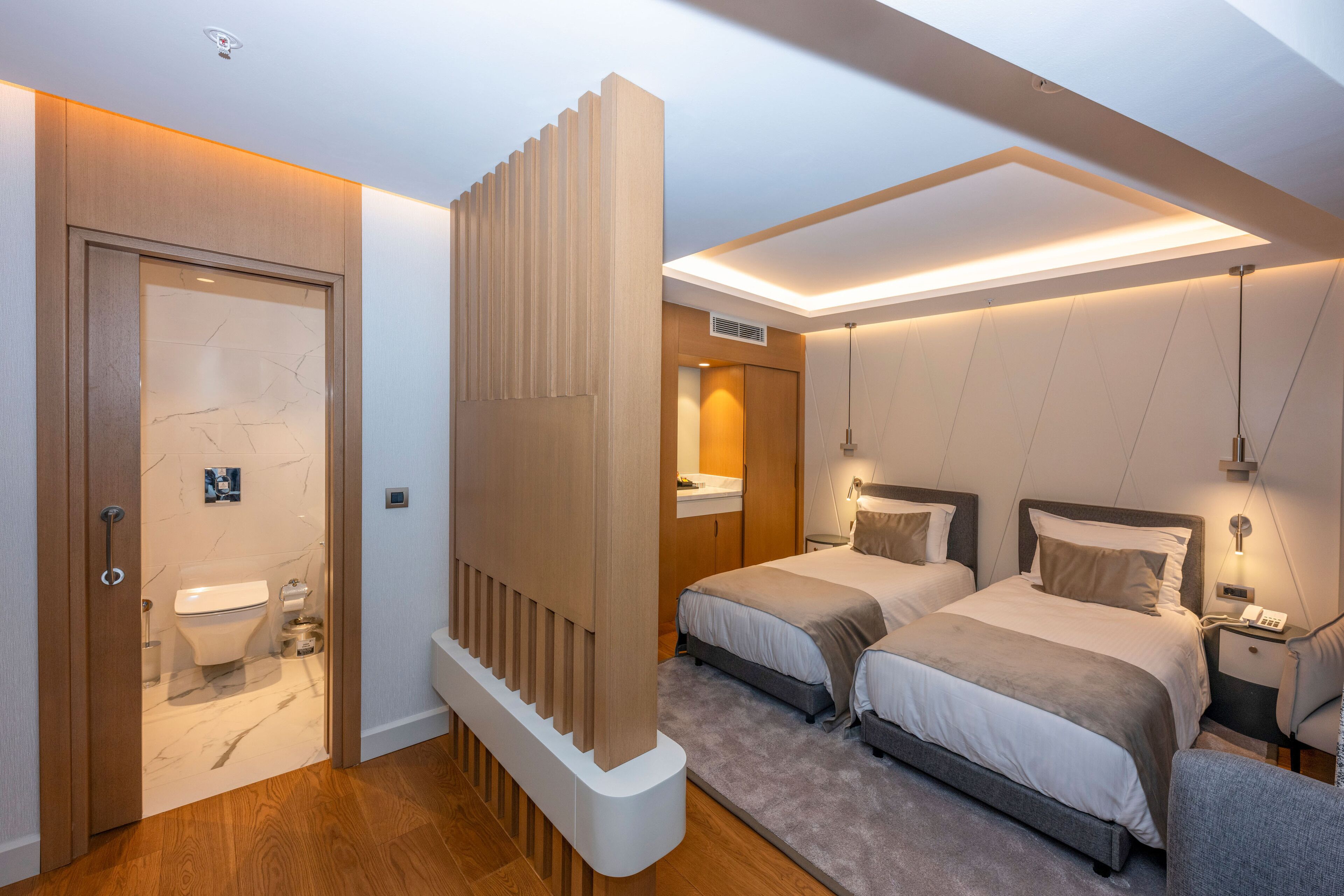 Une chambre d’hôtel moderne avec une porte en bois, une salle de bain, deux lits, une table de chevet et un luminaire au plafond.