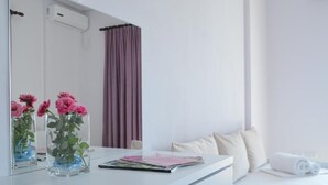 Habitación de hotel con un escritorio blanco, dos jarrones transparentes con flores rosas, una cama con ropa blanca y una cortina morada.