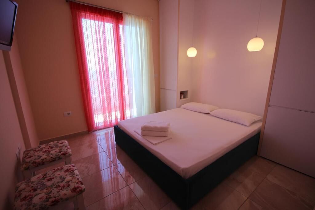A bedroom with a bed, two floral-patterned chairs, a window with red curtains, and a wall-mounted light fixture.