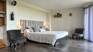 Een slaapkamer met een bed, twee stoelen, een nachtkastje, een lamp, een wandklok, een plank met decoratieve objecten, en een brandblusser.