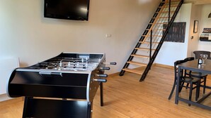 A room with a foosball table, a TV mounted on the wall, a wooden staircase, and a dining area with a table and chairs. - Gîte "La Cordonnerie (MONTGAILLARD LAURAGAIS)