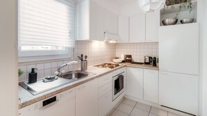 A modern kitchen with white cabinets, a stainless steel sink, a built-in oven, and a window with blinds. - Pink Flat  (Chiasso)