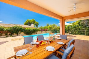 A covered outdoor dining area with a pool, wooden table, chairs, and a ceiling fan.